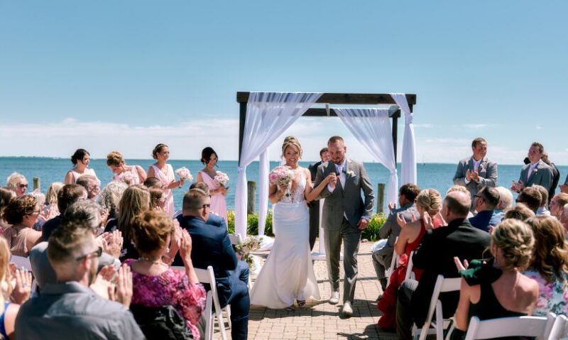 Bride in a fitted white gown with a long train smiles at the camera while holding hands with the groom in a navy suit, surrounded by garden greenery.