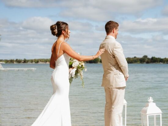 A lakeside first look captures a bride reaching out to her groom’s shoulder on a wooden dock. White lanterns frame the scene against the calm water.
