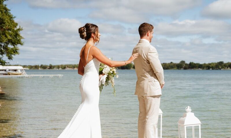 A lakeside first look captures a bride reaching out to her groom’s shoulder on a wooden dock. White lanterns frame the scene against the calm water.