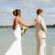 A lakeside first look captures a bride reaching out to her groom’s shoulder on a wooden dock. White lanterns frame the scene against the calm water.