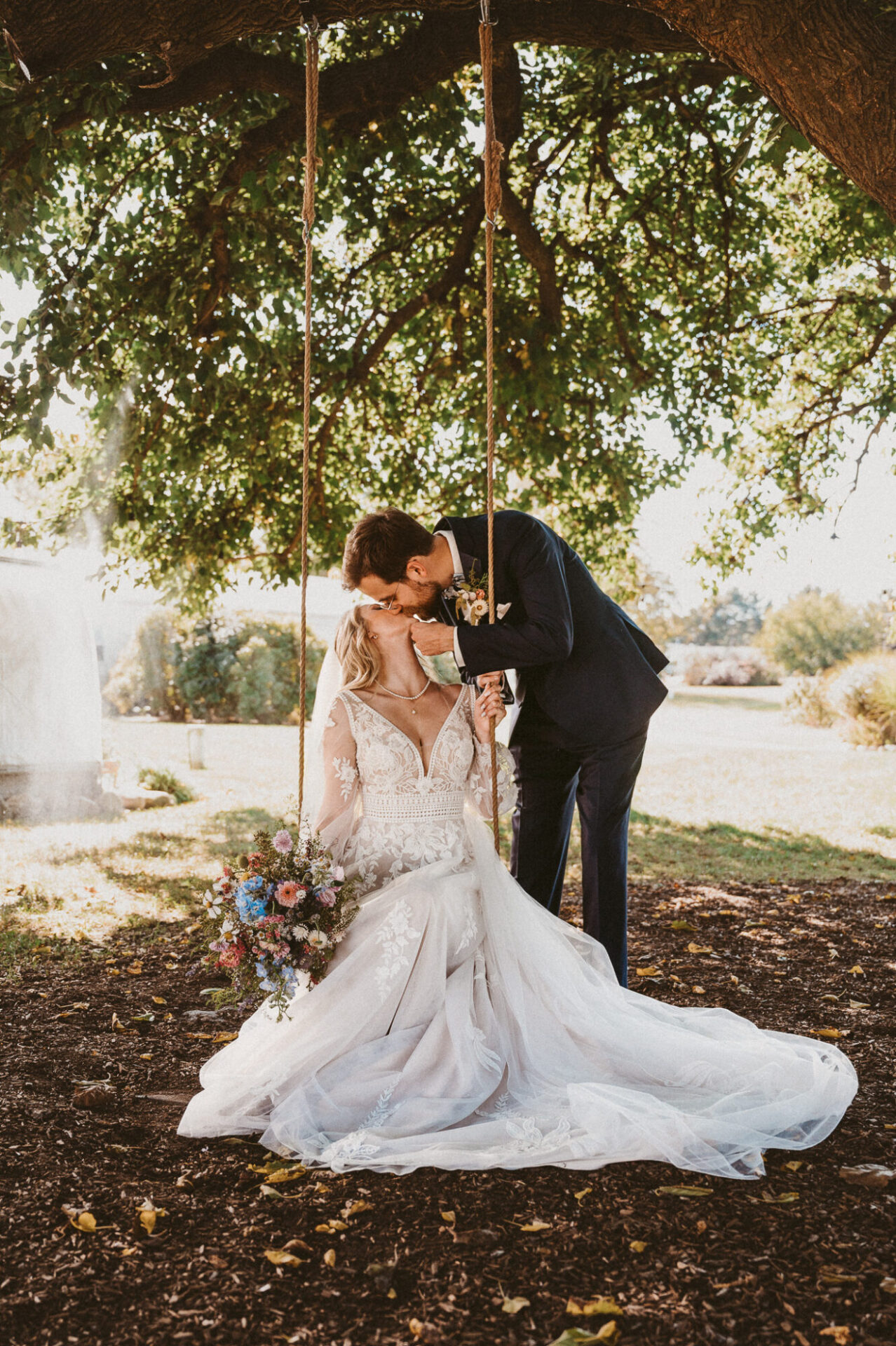 Bride sitting on a hanging swing in a tree while her groom kisses her from behind.