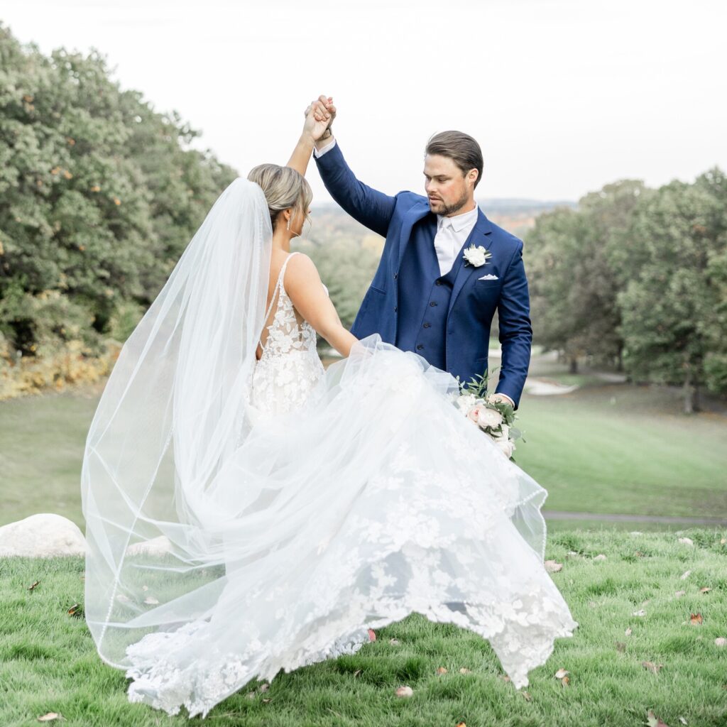 Groom in blue suit spinning his bride in front of him. His bride is twirling and holding her long wedding dress and viel.