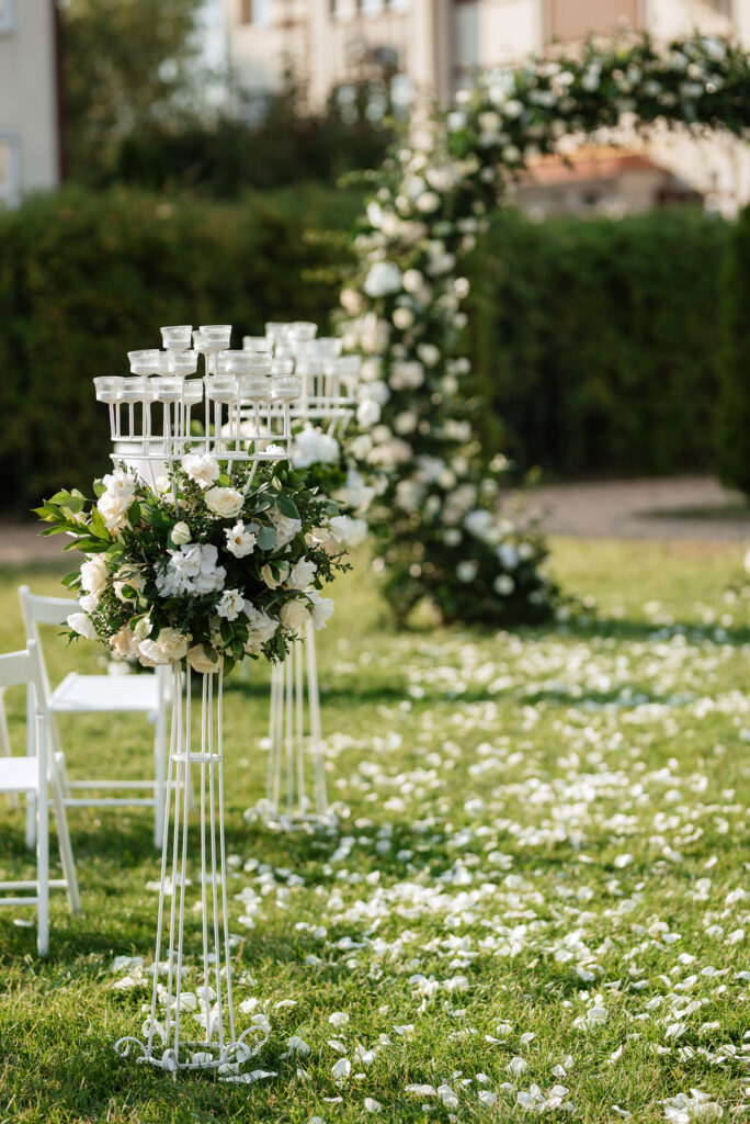 Elegant white floral arrangements displayed on tall stands lining the aisle, with white rose petals scattered across the green grass and a floral arch in the background.