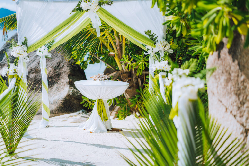 Tropical beach wedding ceremony setup with white and green draped fabric, lush greenery, and a round table decorated with seashells on the sandy shore.