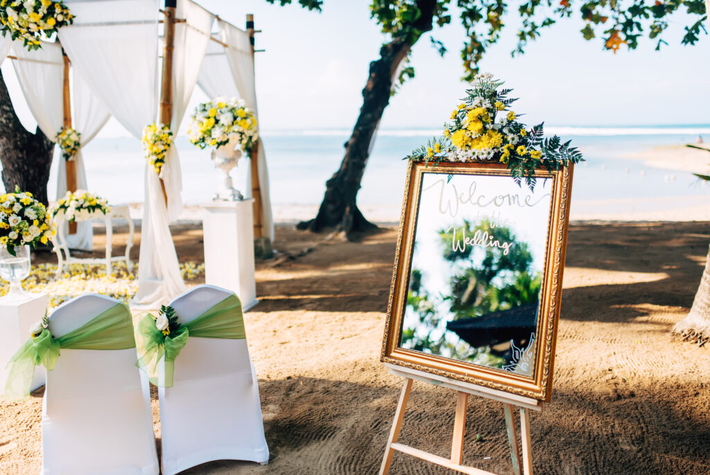 Beachfront wedding setup featuring two white chairs decorated with green sashes, floral arrangements, and a gold-framed mirror sign welcoming guests to the wedding.