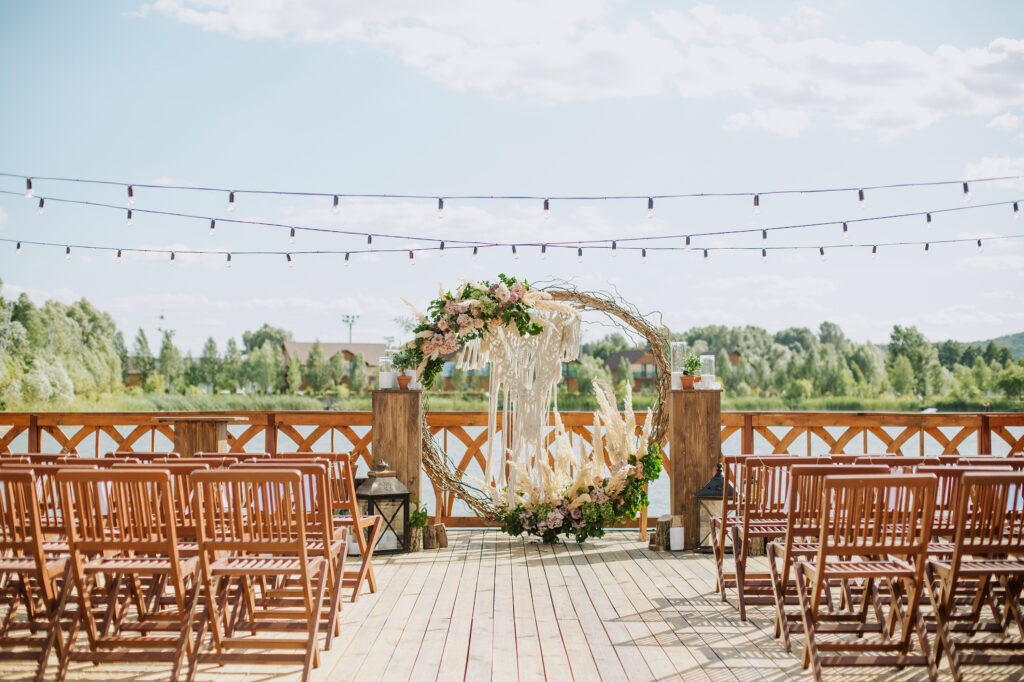 A rustic lakeside wedding ceremony setup featuring wooden chairs, string lights, and a large circular floral arch with pampas grass and blush flowers.