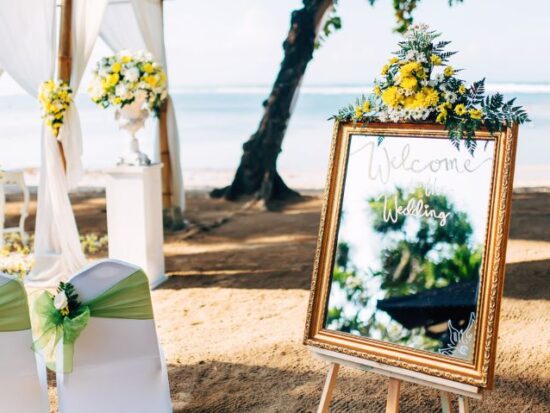Beachfront wedding setup featuring two white chairs decorated with green sashes, floral arrangements, and a gold-framed mirror sign welcoming guests to the wedding.