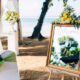Beachfront wedding setup featuring two white chairs decorated with green sashes, floral arrangements, and a gold-framed mirror sign welcoming guests to the wedding.