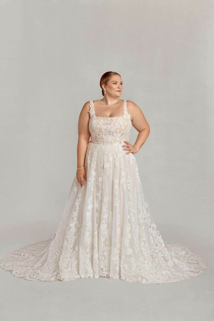 A plus-size bride stands confidently in a romantic A-line lace wedding dress with wide straps and floral appliqués, set against a plain gray backdrop.