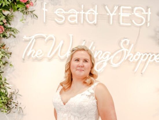 A bride wearing a sleeveless floral lace A-line gown stands in front of The Wedding Shoppe’s “I said YES” neon sign surrounded by floral arrangements.