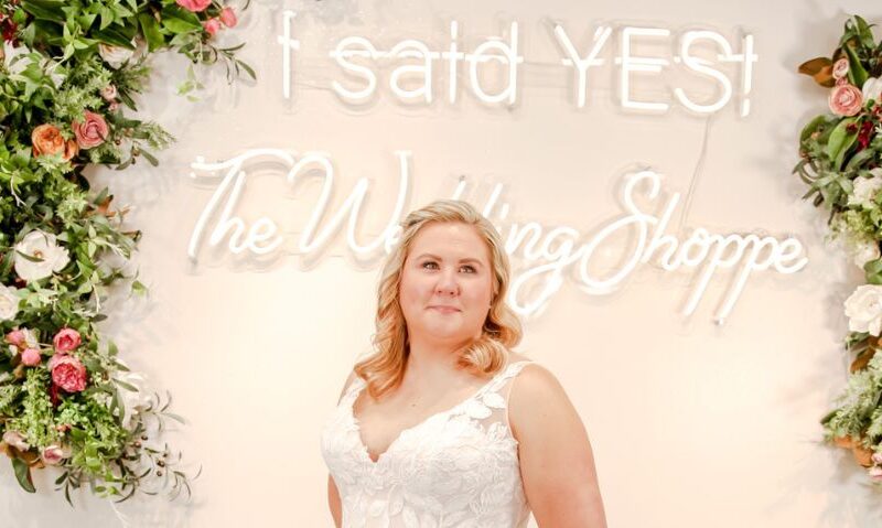 A bride wearing a sleeveless floral lace A-line gown stands in front of The Wedding Shoppe’s “I said YES” neon sign surrounded by floral arrangements.