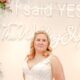 A bride wearing a sleeveless floral lace A-line gown stands in front of The Wedding Shoppe’s “I said YES” neon sign surrounded by floral arrangements.