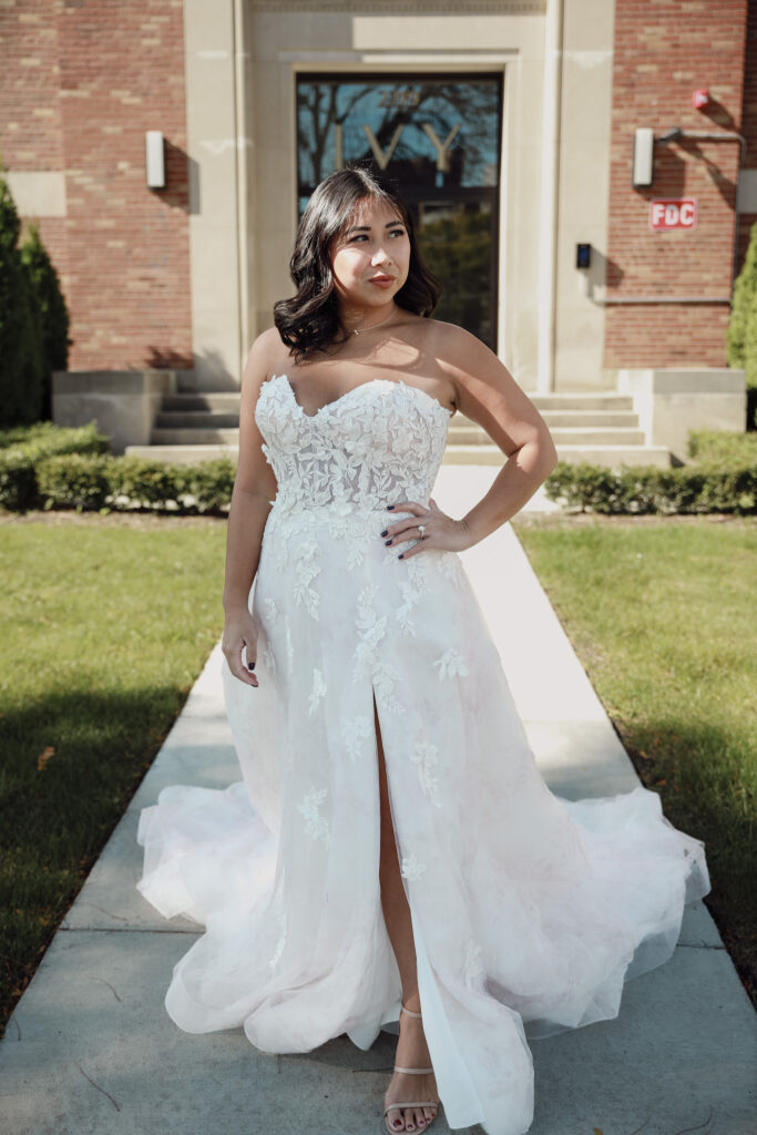 A bride stands confidently outdoors in front of a brick building, wearing a strapless white lace wedding dress with a sweetheart neckline and a high slit.