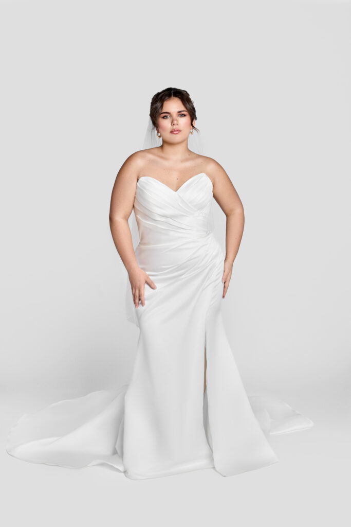 A plus-size bride stands in a fitted strapless white wedding gown with a sweetheart neckline and flowing train, set against a plain gray background.