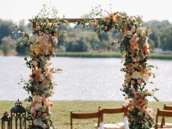 A beautiful outdoor wedding ceremony arch decorated with peach, blush, and white flowers, along with green foliage, positioned by a lake with a lantern and table setup nearby.