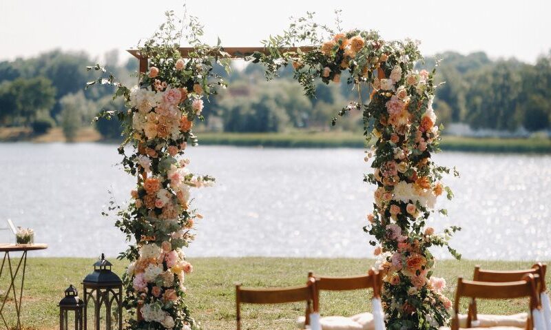 A beautiful outdoor wedding ceremony arch decorated with peach, blush, and white flowers, along with green foliage, positioned by a lake with a lantern and table setup nearby.