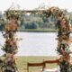 A beautiful outdoor wedding ceremony arch decorated with peach, blush, and white flowers, along with green foliage, positioned by a lake with a lantern and table setup nearby.