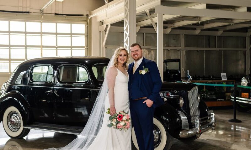 Bride and groom posing beside a vintage black car in a bright, industrial-style venue, with the bride in a white wedding dress and the groom in a navy-blue suit.