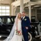 Bride and groom posing beside a vintage black car in a bright, industrial-style venue, with the bride in a white wedding dress and the groom in a navy-blue suit.
