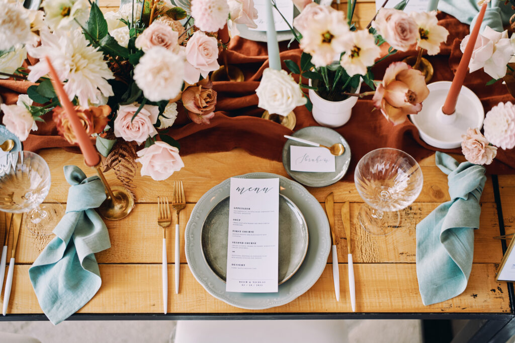 Stylish wedding tablescape featuring muted orange, blush, and ivory florals, teal napkins, gold flatware, and a printed menu on a rustic wooden table.