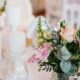 Romantic wedding reception table with a pastel floral centerpiece of blush pink roses, white blooms, greenery, and soft candlelight in the background.