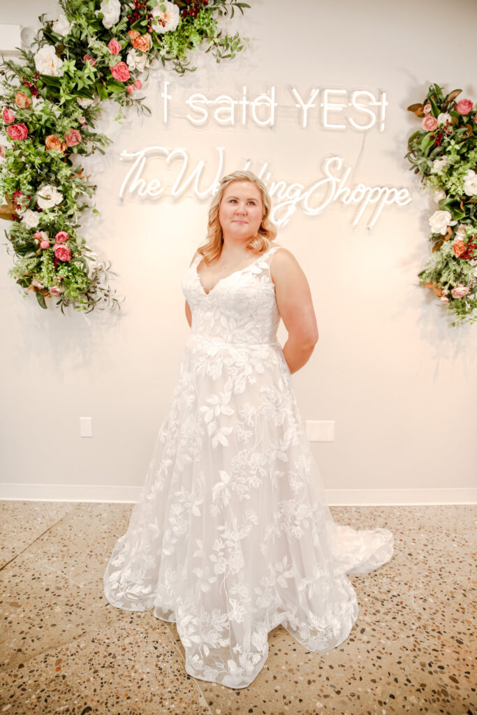 A plus-size bride smiles while standing in a bridal boutique in front of a neon sign reading “I said YES! The Wedding Shoppe,” wearing a sleeveless lace A-line wedding dress with floral detailing.
