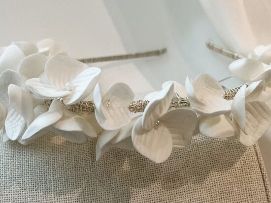 Close-up side view of a bridal headband wrapped in delicate white flowers with petal textures, displayed on a neutral fabric stand.