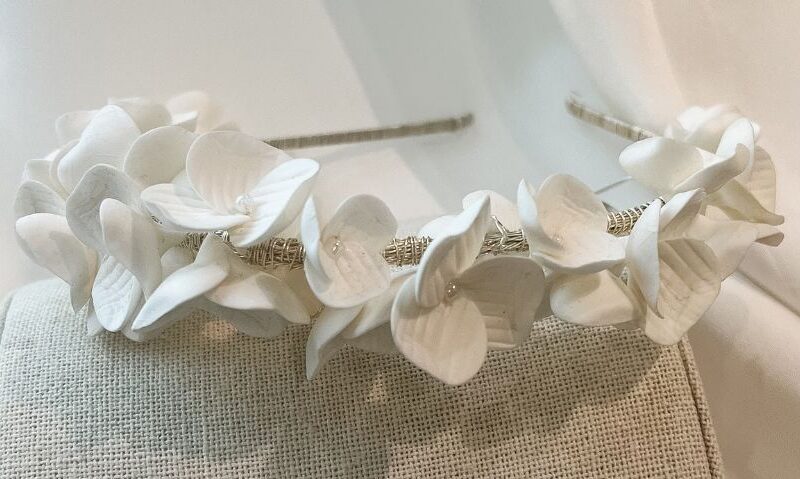 Close-up side view of a bridal headband wrapped in delicate white flowers with petal textures, displayed on a neutral fabric stand.