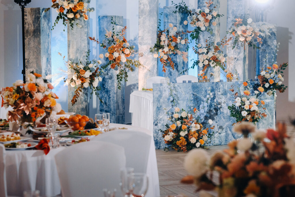 Elegant wedding reception setup with a blue marble sweetheart table backdrop decorated with orange, white, and peach floral arrangements.