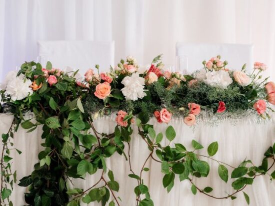 A romantic sweetheart table decorated with lush greenery and pink, peach, and white florals, accented with pillar candles on each side.