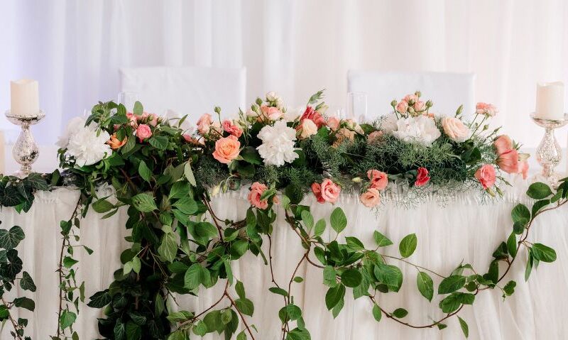 A romantic sweetheart table decorated with lush greenery and pink, peach, and white florals, accented with pillar candles on each side.