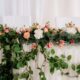 A romantic sweetheart table decorated with lush greenery and pink, peach, and white florals, accented with pillar candles on each side.