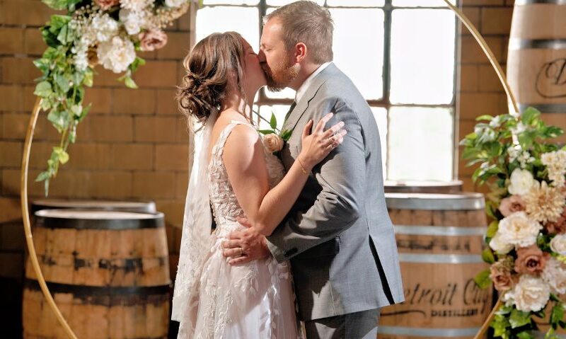 A newlywed couple shares a kiss in front of a circular floral arch, with wooden whiskey barrels and exposed brick walls adding to the cozy, industrial-chic ambiance.