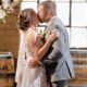 A newlywed couple shares a kiss in front of a circular floral arch, with wooden whiskey barrels and exposed brick walls adding to the cozy, industrial-chic ambiance.