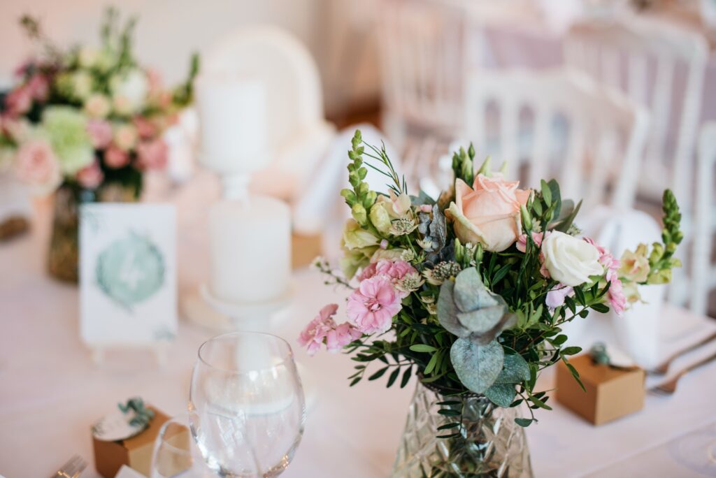 Close-up of a romantic wedding centerpiece featuring blush pink roses, white flowers, and greenery in a glass vase on a table with candles and glassware.