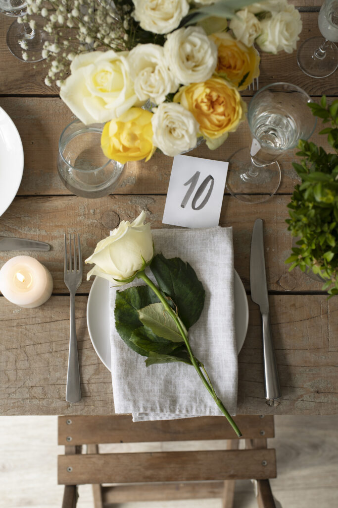 Rustic wedding table with a white rose placed on a folded napkin, surrounded by yellow and white flowers, glassware, and a small table number card.