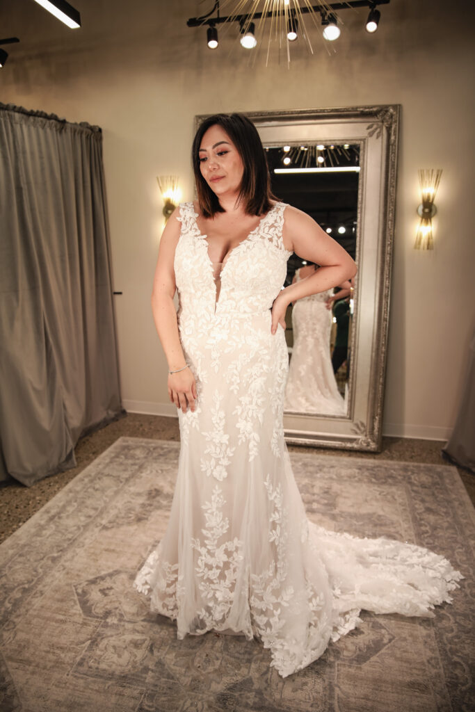 A bride models a sleeveless fitted wedding gown with floral lace detailing and a plunging neckline in a bridal boutique fitting room.