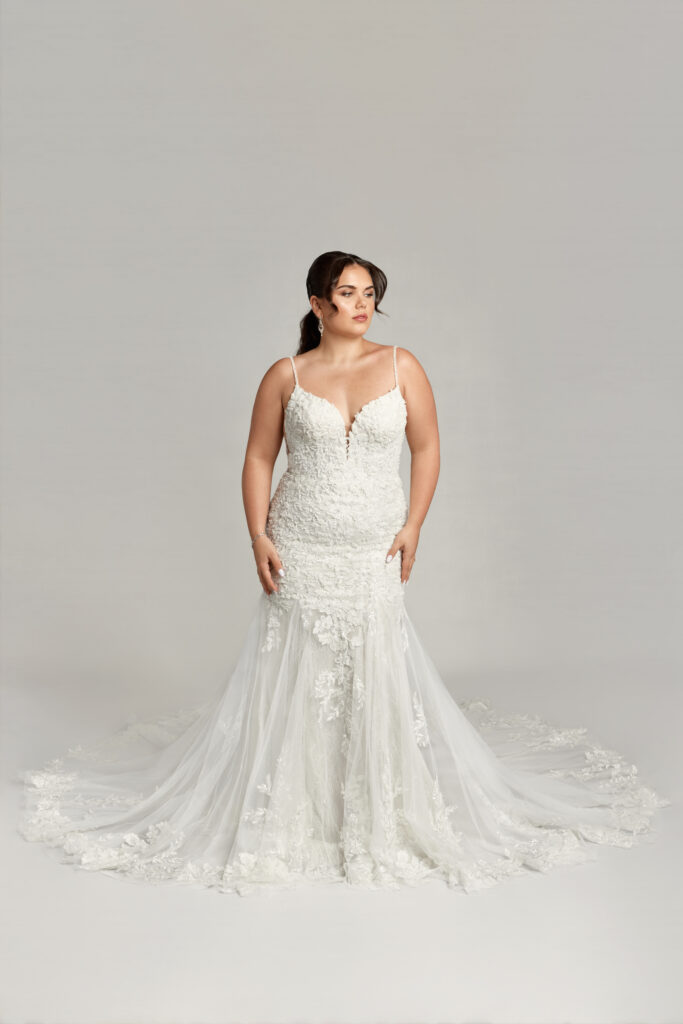 A plus-size bride poses in a fitted mermaid-style wedding dress with thin straps, floral lace appliqués, and a dramatic train against a plain gray background.