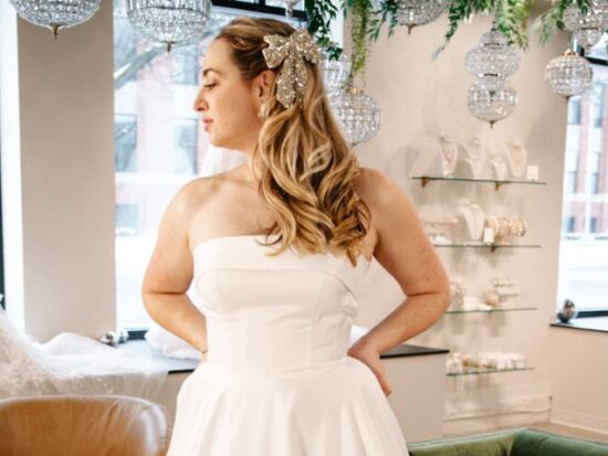 A bride poses in a strapless ballgown inside a boutique, showing the soft structure of the bodice and the full gathered skirt.
