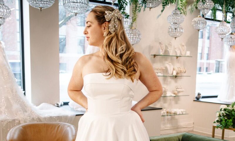 A bride poses in a strapless ballgown inside a boutique, showing the soft structure of the bodice and the full gathered skirt.