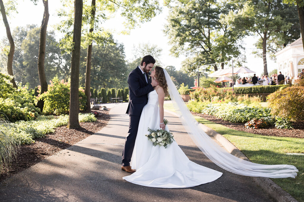 Bride and groom embracing on a landscaped path, bride in a white dress with long train and veil holding a white bouquet.
