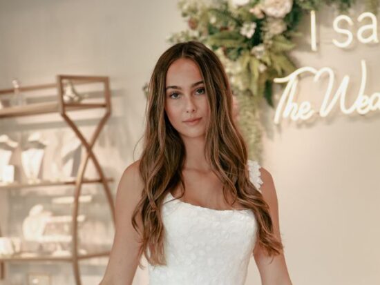 Bride standing in front of “I said Yes” sign at The Wedding Shoppe, wearing a floral lace gown with long, loose curls.