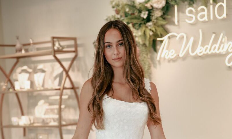 Bride standing in front of “I said Yes” sign at The Wedding Shoppe, wearing a floral lace gown with long, loose curls.
