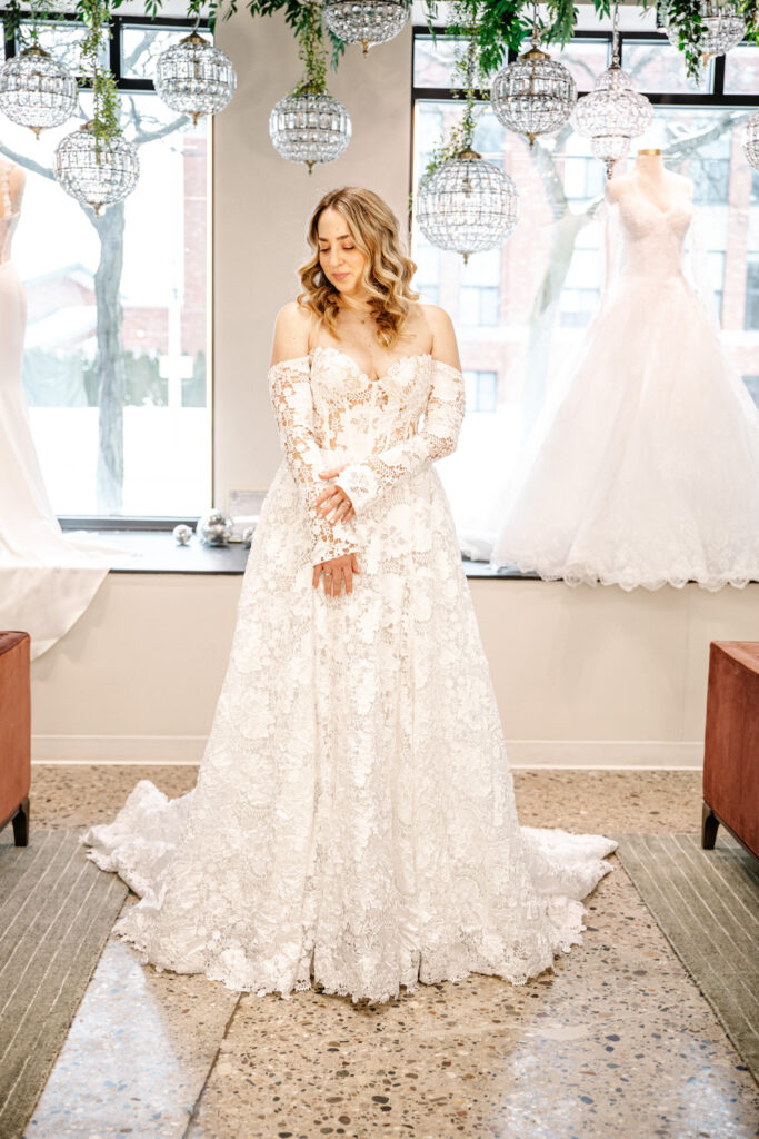 Bride in an off-the-shoulder lace wedding dress with long sleeves, standing beneath crystal chandeliers near a window.