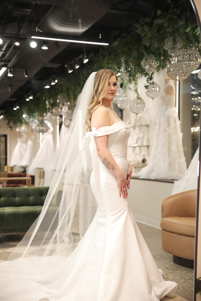 Bride wearing a sleek, off-the-shoulder satin mermaid gown with a long veil and bow detail in a modern bridal salon.