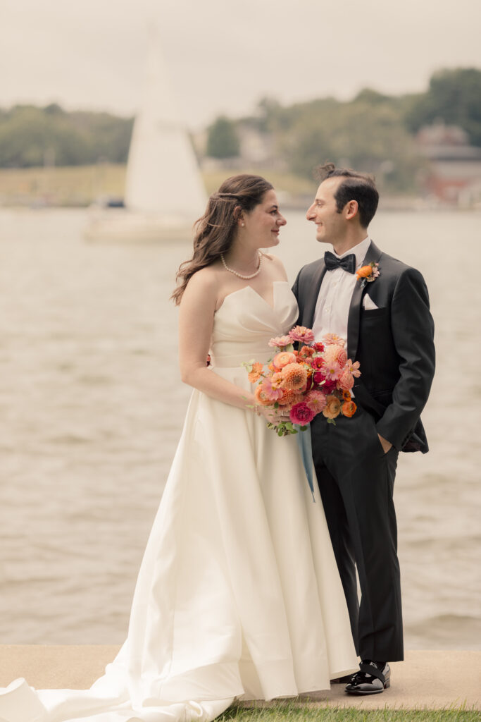 Bride and groom standing beside a lake, bride wearing a strapless gown and holding a colorful bouquet of orange and pink flowers.