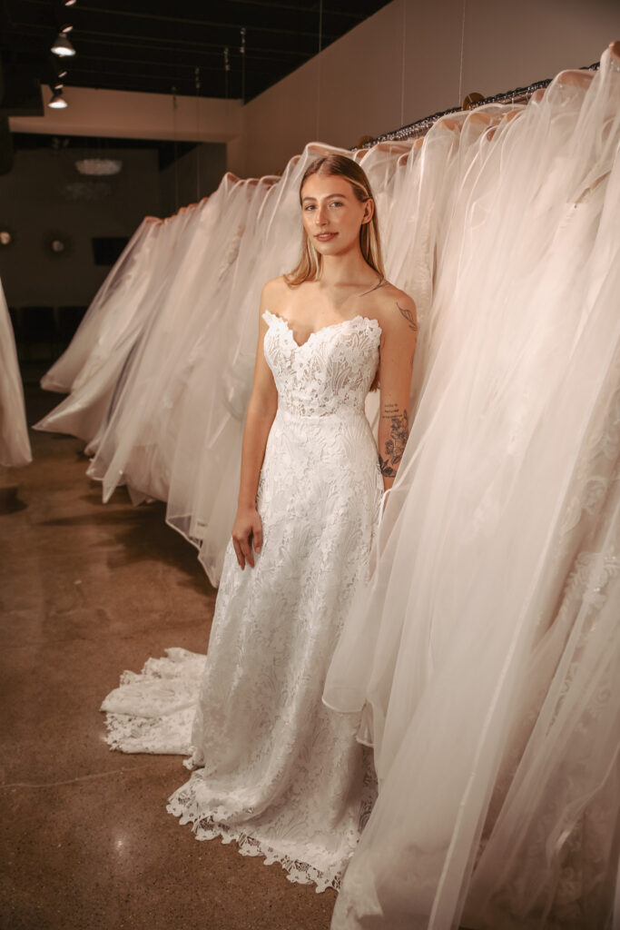 The same bride in the lace gown stands by racks of wedding dresses, showcasing the floral lace detail and scalloped train.