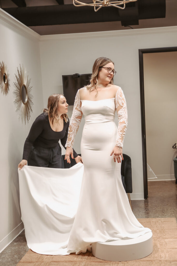 A bride wearing a long sleeve wedding gown with a fitted silhouette stands on a pedestal while a stylist adjusts the train.