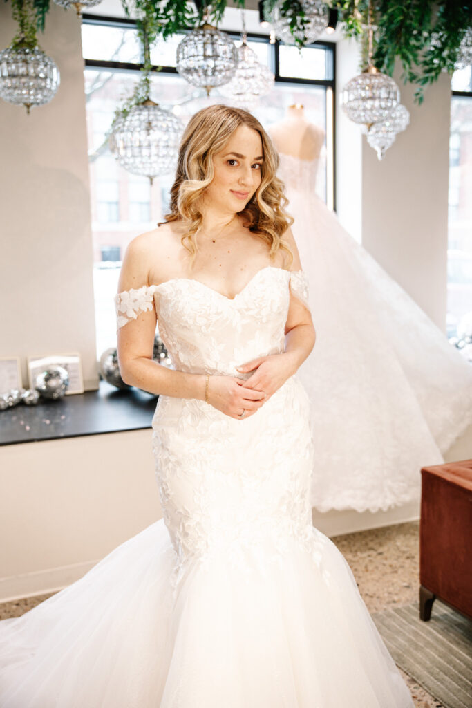 A bride with soft curls wears an off-the-shoulder mermaid gown with floral appliqués and a voluminous tulle skirt in a bright bridal shop.