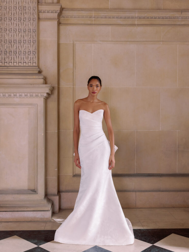 Bride wearing a strapless white fit-and-flare gown in a grand hall with decorative molding and a tiled floor.